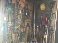 View of multiple garden tools hanging on a wooden wall, including shovels, stirrups, and other farming implements.
