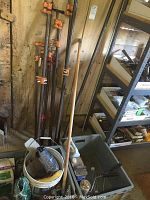Photo of several long metal bar clamps leaning against a wall, showing orange and red clamp heads and wooden handles.