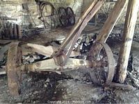 Antique wagon wheel attached to an axle with wooden and metal parts, placed on a barn floor with stone wall background.