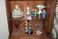 Display cabinet with German porcelain seated lady figurine, 3 ceramic Asian figures, decorative bowl, glass collectibles, and other items.