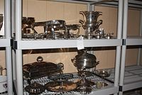 Multiple silver-plated serving pieces displayed on shelving including chafing sets and trays