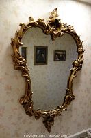 Full view of the gilded Rococo style wood wall mirror hanging on a wallpapered wall with framed pictures reflected in the glass.