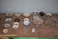 Wide shot showing entire assortment of collectable dishes including porcelain, crystal, and carnival glass pieces laid out on carpeted floor.