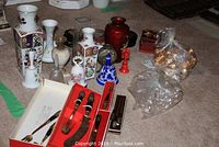 Photo of multiple ceramic and porcelain vases, serving utensil set in red box, decorative items including napkin rings and bells displayed on carpeted floor.