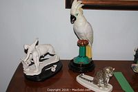 Wooden surface holding three figurines: unglazed white bull and matador, porcelain parrot on pedestal, and hand-painted spotted big cat.