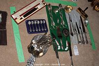 Wide view of assorted silver-plated serving utensils including tongs, spoons, forks, carving knives, a boxed Royal Crown Derby knife, and a set of six small teaspoons in a blue case.