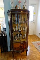 Full view of curved-front vitrine showing mahogany finish, brass mounts, glass shelves, lithograph base panel and decorative top gallery.