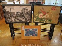 Three sets of decorative tray table tops with different designs arranged with two on stands and one laying on floor.