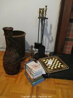Photo showing entire lot including tall carved wood vase, fireplace tool set, statuette, game board, and stack of books on wooden floor near fireplace.