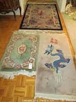 Three area rugs arranged on hardwood floor: largest navy-bordered landscape design, medium green floral medallion rug, and beige rug with blue dragon motif