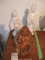 Three figurines pictured together on wood surface: two white porcelain Japanese women figurines and one colorful ceramic Hotei figure in front.