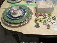Full view of stacked decorative plates, demitasse cup and saucer, and miniature porcelain flowers spread on table, some flowers near a box with tissue paper.