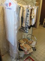 Photo showing multiple tablecloths and runners hanging on hangers alongside a cart piled with folded linens such as napkins and smaller table covers.
