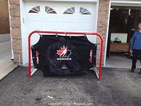 Front view of the hockey net with attached black Aim Pro shooting target featuring a maple leaf and Canada logo. Red steel frame and white netting visible.