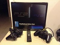 PlayStation 2 console black box with network adapter pack shown, two wired controllers and remote control in front.
