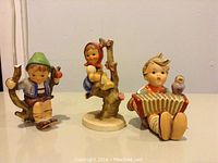 Three Goebel Hummel figurines arranged in a row showing front view, displaying children in different poses with accompaniment of birds and a horn, on a plain background.