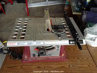 Front view of Delta table saw showing metallic work surface, clear plastic blade guard, and maroon base with control dial.