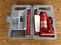 Hydraulic bottle jack and two detachable handles inside gray molded plastic carrying case on wooden floor.