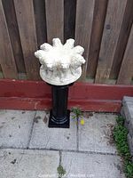 Front view of the concrete lion head bird bath mounted on a black pedestal stand in an outdoor tiled patio area against a wooden fence.