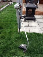 Overall view of the power trimmer leaning against a pole on grass near an outdoor stove.