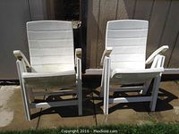 Two white plastic outdoor folding chairs side by side on outdoor concrete surface.