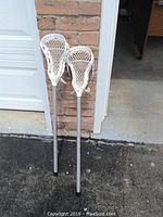 Two white lacrosse paddles leaning against a brick wall outside a garage. The mesh heads are clearly visible, as are the grey shafts.
