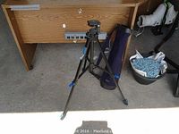 Camera tripod standing on concrete floor near wooden desk with soft fabric carrying case nearby.