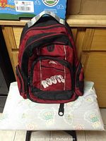 Front view of a red and black Roots teen backpack, showing 'ROOTS' embroidered on the front pocket and multiple compartments.