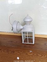 White exterior lantern-style light with motion sensor, showing side profile and base, sitting on wooden surface.