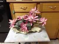 Artificial pink lotus flower plant with multiple blossoms and buds, green leaves, set in an off-white clay log base, shown from front angle on a floral fabric surface against wooden cabinetry.