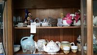 Wide view of two wooden shelves with assorted teapots, glassware, ceramics, and decorative items.