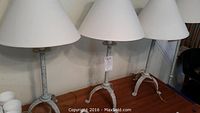 Photo showing three identical tabletop lamps with white fabric cone shades and distressed white metal tripod bases on a wooden surface.