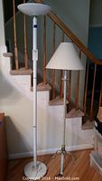 Photo of two floor lamps standing by staircase, white halogen torchiere style on left, traditional metal lamp with fabric shade on right.