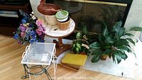 Small wooden round table, white metal mesh plant stand, green metal plant stand, large leafy green plant in a wicker basket, variegated plant, artificial flowers, duck-shaped wicker basket, assorted ceramic and plastic planters.