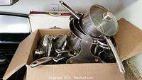 Full view of assorted stainless steel pots and pans piled in a box, some with glass lids and some without.