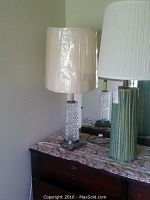 Photo showing the two lamps side by side on a dresser with floral cloth. One lamp has a clear pressed glass base on a metal stand with a beige shade, the other has a green vertically grooved ceramic base with a white pleated shade.