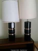 Pair of black cylindrical table lamps with silver accent bands on the bases, each topped with a mismatched white lampshade. Lamps are approximately 34 inches tall, placed on a wooden surface.