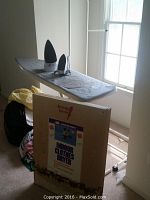 Gray ironing board with padded cover, two irons placed on it, assorted linens and a boxed indoor clothes dryer visible on floor nearby.