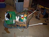 Overall view of wheelbarrow containing cleaning supplies, garden hose, and cardboard box with cleaning liquids.
