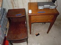 Photo showing both the wooden side table with multiple tiers and the sewing table with drawers, placed against a wall and surrounded by boxes.