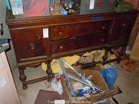 Full view of vintage wooden dresser with carved side cabinets, two center drawers, and turned legs. Various items on top and some clutter below.