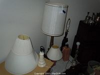 Six assorted table lamps with visible shades; two shades show fraying or dents; lamps shown on table and floor near wall.