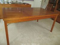Front angle view of the table showing warm teak wood finish and straight legs on carpeted floor.