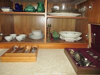 View of wooden cabinet shelving showing white ceramic teacups and saucers, green Christmas-themed ceramic pieces, glass serving plates and bowls, brass candlestick holders, vegetable serving trays, and two silverware sets in velvet-lined cases.