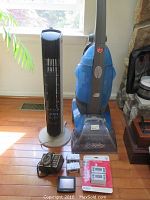 Blue Hoover Agility rug cleaner, black and white tower fan, two black electric timers, small digital clock/timer, and packaged white digital timer on wooden floor near window.