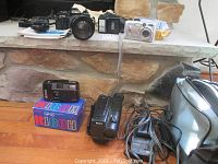 Photo showing Minolta X-700 camera with lens, Sony Handycam, Kodak EasyShare camera, disposable Ricoh cameras and soft carrying case on floor in front of stone fireplace.