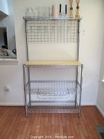 Full view of metal bakers rack against wall with wooden butcher block top, wire shelves, and back grid panel with hooks.