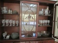 Wide view of the full collection displayed on wooden shelves in a china cabinet, showing stemware goblets, tumblers, bowls, pitchers, salt and pepper shakers, and glass plates.