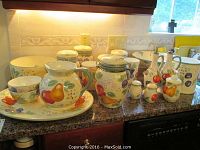 Wide view of the entire set of porcelain kitchenware arranged on the kitchen counter, showing canisters, bowls, pitcher, cruets, and serving platters, all with lively fruit decorations in yellow, red, purple, and green colors.