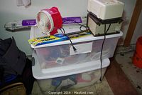 Various household items in stacked clear plastic storage containers, including cords, a red and white fan, and a packaged blind visible on top.